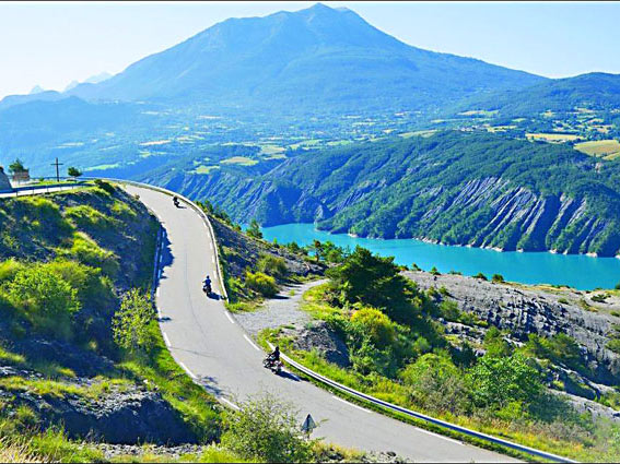 Photo d'une route avec 3 motos suplombant un lac aux eaux bleu turquoise dans les Alpes
